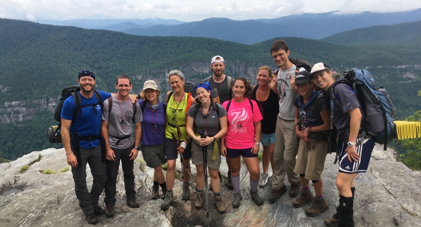 A group of people pose for a photo on a rocky overlook, with the blue ridge mountains below. 